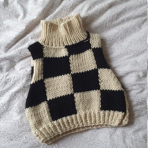 Może przedstawiać: Czarno-biała w kratę dzianinowa kamizelka z golfem. Kamizelka wykonana jest z grubego włóczki i ma luźny krój.