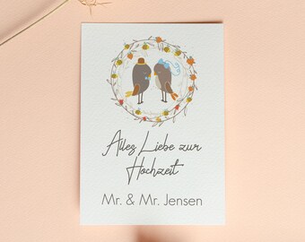 May include: A white card with a floral wreath and two birds inside. The birds are wearing hats and a bow. The text on the card reads "Alles Liebe zur Hochzeit" and "Mr. & Mr. Jensen".
