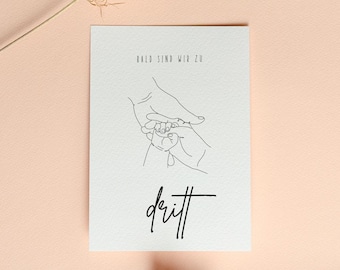 May include: A white card featuring a line drawing of hands cradling a baby's hands, with the words "bald sind wir zu dritt" in black script. The card is on a peach background with dried flowers.