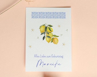 May include: A white greeting card with a blue floral border and a watercolor illustration of lemons. The card has the text "Alles Liebe zum Geburtstag Mareike" in blue script. Dried flowers are in the foreground.