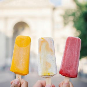 May include: Three frozen fruit popsicles on wooden sticks held up in a row. The left popsicle is solid bright orange, the center one is a creamy white with orange swirls, and the right popsicle is a solid deep red color, all with a smooth texture.