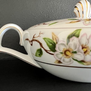 May include: White porcelain serving dish with a gold trim and a floral design featuring white and pink flowers with green leaves. The dish has a lid with a gold knob.