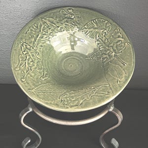 May include: A green ceramic bowl with embossed leaf designs, resting on a decorative metal stand. The bowl has a glossy finish and a shallow, rounded shape. The metal stand has a curved, scroll-like design.