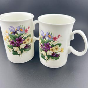 May include: Two white ceramic mugs with floral designs. The mugs feature a bouquet of purple, yellow, and white flowers with green leaves.