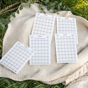 May include: A collection of white saving challenge cards, each featuring a grid of circles representing £5, are displayed on a cream knitted blanket. The cards are labelled "£250 Saving Challenge."