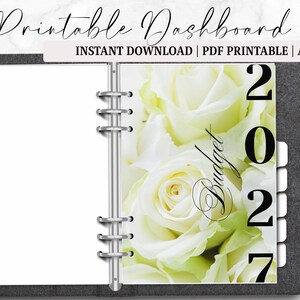 May include: A gray A6 planner with a floral design and the text "Printable Dashboard". The right page features white roses and the year "2027" with the word "Budget". The planner is open, revealing blank pages.