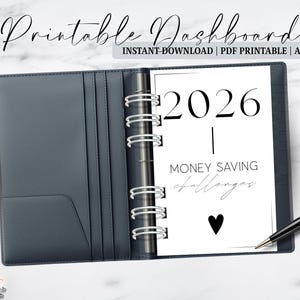 Puede incluir: Un archivador A6 gris oscuro con una inserción blanca que muestra el año 2026 y el texto "Money Saving Challenges". El archivador está abierto, revelando la inserción y un mecanismo de anillas plateado. Un bolígrafo negro descansa a la derecha.