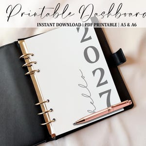May include: A black binder with gold-coloured rings holds a white page with the year "2027" printed in grey. A rose gold pen rests on the page. The text "Printable Dashboard" and "INSTANT DOWNLOAD | PDF PRINTABLE | A5 & A6" are at the top.