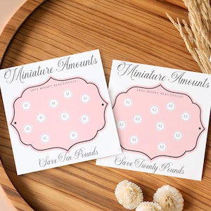 Könnte beinhalten: Zwei quadratische weiße Spar-Tracker aus Papier mit rosafarbenen, geschwungenen Rahmen. Die linke Karte zeigt den Text Miniature Amounts, Save Money Beautifully und Save Ten Pounds mit £1-Kreisen. Die rechte Karte zeigt Save Twenty Pounds mit £2-Kreisen.