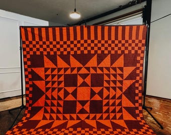 May include: A large, handmade quilt in shades of orange and burgundy. The quilt features a geometric pattern with star and square designs. The top and bottom borders have a striped pattern, while the center showcases a complex arrangement of shapes.