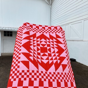 Könnte beinhalten: Eine große, handgefertigte Steppdecke mit einem auffälligen geometrischen Design. Die Steppdecke weist ein sich wiederholendes Muster aus roten und rosa Quadraten und Dreiecken auf, das einen kühnen visuellen Effekt erzeugt. Die Steppdecke wird im Freien ausgestellt.