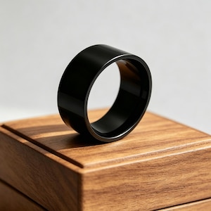 Puede incluir: Un anillo negro sólido con un acabado liso y pulido y un diseño de banda ancha. El anillo presenta una forma circular con un grosor constante, mostrando un color negro profundo, de mate a satinado, que refleja la luz en sus bordes curvos.
