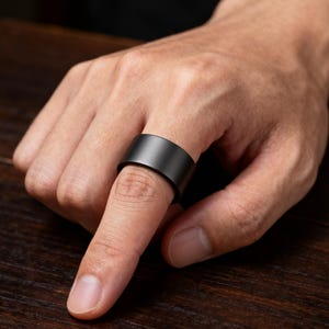 以下が含まれることがあります： 幅広のバンドが付いたマットブラックのリングが指に装着されています。 リングは滑らかでしっかりとした表面とミニマリストなデザインです。 リングは濃いグレー色で、金属製のように見えます。 背景は濃い木製の表面です。
