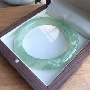Op de afbeelding: Een gladde, doorschijnende lichtgroene jade armband met subtiele witte, wolkachtige insluitingen. De ronde armband wordt gepresenteerd in een donkerbruine houten displaydoos met een crèmekleurige voering en een zachte, gevoerde basis.