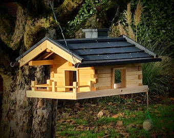 May include: A wooden bird feeder shaped like a small cabin with light natural wood walls, a dark gray shingled roof, and a chimney. The structure features a front porch with a railing, open side entrances, and a hanging mechanism for outdoor use.