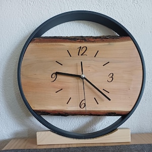 Könnte beinhalten: Eine runde Wanduhr mit einer rechteckigen, hellen Holzplatte mit natürlicher Baumkanten-Optik als Zifferblatt. Die Uhr hat schwarze Zeiger und schwarze Ziffern für 3, 6, 9 und 12. Sie ist in einem dünnen schwarzen Ring auf einem Holzsockel.