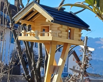 Könnte beinhalten: Ein Vogelhaus aus Holz mit schwarzem Dach, das einer kleinen Hütte ähnelt, ist auf einem Holzständer montiert. Das Vogelhaus hat eine kleine Veranda und Fenster. Die Struktur befindet sich im Freien vor dem Hintergrund von Bäumen und blauem Himmel.