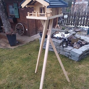 Könnte beinhalten: Ein Vogelhaus aus Holz mit dunklem Dach und kleinem Balkon, montiert auf einem dreibeinigen Ständer. Das Vogelhaus ist aus hellem Holz gefertigt und hat einen Schornstein. Der Ständer ist ebenfalls aus hellem Holz gefertigt und steht auf einem Rasen.