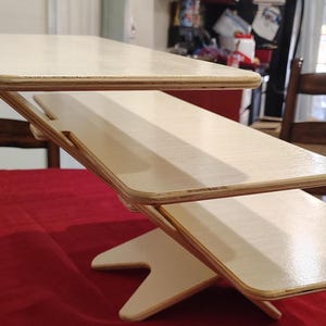 May include: A light-colored, wooden, two-tiered desk organizer with a unique angled design. The desk organizer is made of light-colored wood and has a natural finish. The desk organizer is on a red surface.