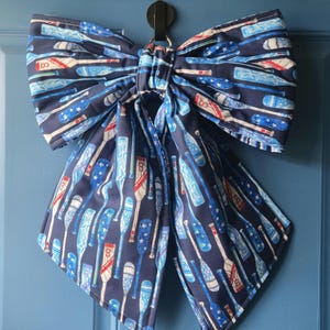 May include: A large fabric bow featuring a dark blue background with a repeating pattern of baseball bats in shades of blue, white, and red. The bats are decorated with stars and stripes, and the bow is suspended from a black metal hook.