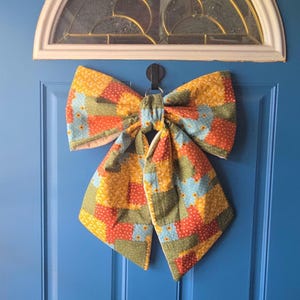 Op de afbeelding: Een grote stoffen strik met een patchworkontwerp met vierkanten in tinten geel, oranje, groen en lichtblauw. De stof heeft overal een klein bloemen- en stippenpatroon. De strik is geconstrueerd met een gewatteerde textuur en een centrale knoop.