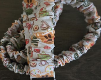 Puede incluir: Un cordón de tela con un estampado repetido de hombrecitos de jengibre, galletas y otras golosinas festivas. El cordón es blanco con detalles en naranja, rojo, verde y marrón. Tiene un cierre metálico y un llavero plateado.