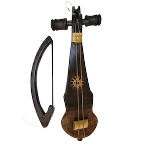 Op de afbeelding: Een donkerbruin, handgemaakt snaarinstrument met een strijkstok. Het instrument heeft een unieke vorm, met een decoratief cirkelvormig ontwerp en drie snaren. De strijkstok is gebogen en gemaakt van hetzelfde donkere hout. Het is waarschijnlijk een volksinstrument.