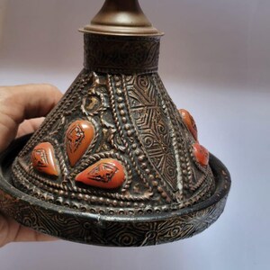 Op de afbeelding: Een decoratief metalen deksel met een conische vorm en ingewikkelde houtsnijwerk. Het deksel is versierd met oranje edelstenen en een messing knop.
