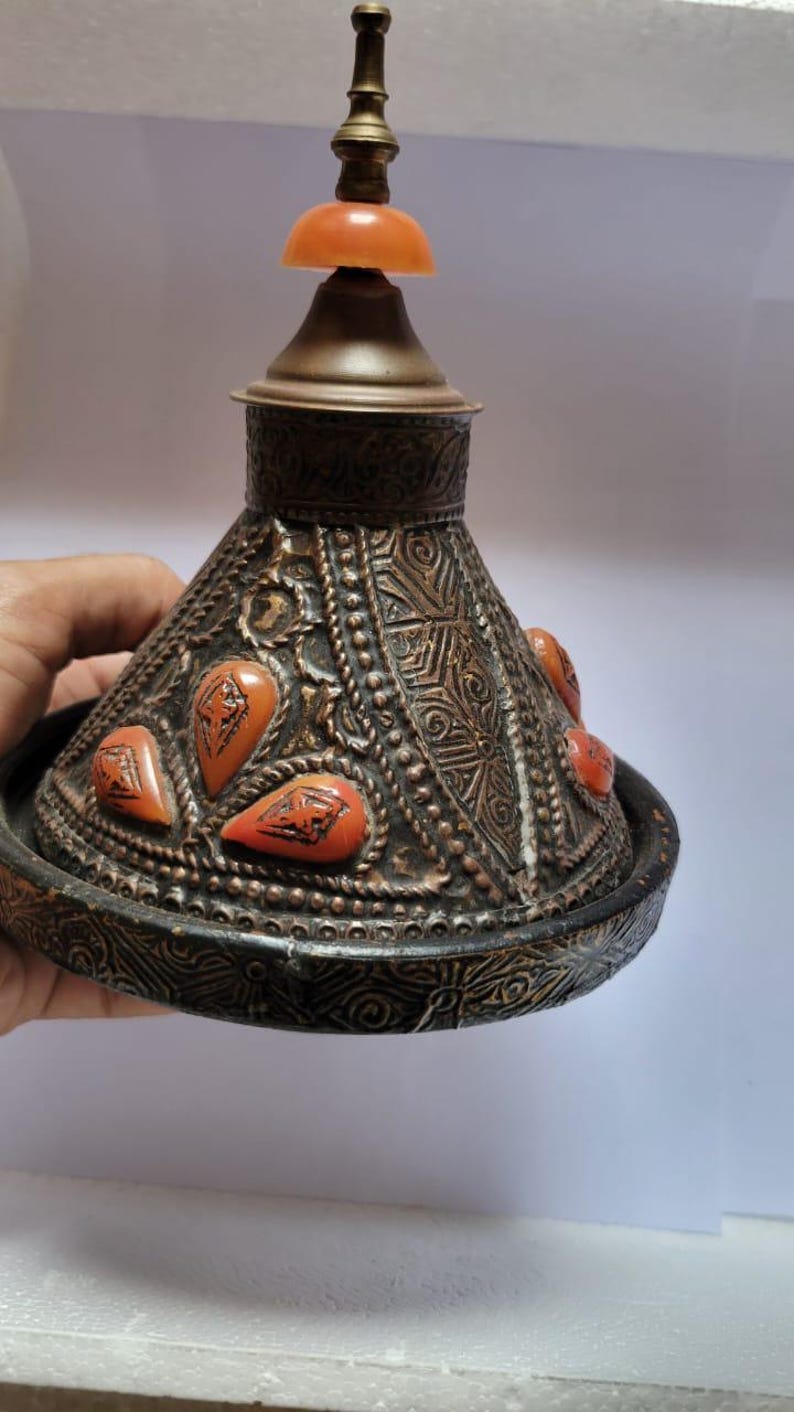 Op de afbeelding: Een decoratief metalen deksel met een conische vorm en ingewikkelde houtsnijwerk. Het deksel is versierd met oranje edelstenen en een messing knop.