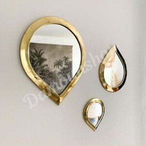 May include: Three gold teardrop-shaped mirrors of varying sizes hanging on a white wall. The largest mirror is in the centre and the two smaller mirrors are on either side.