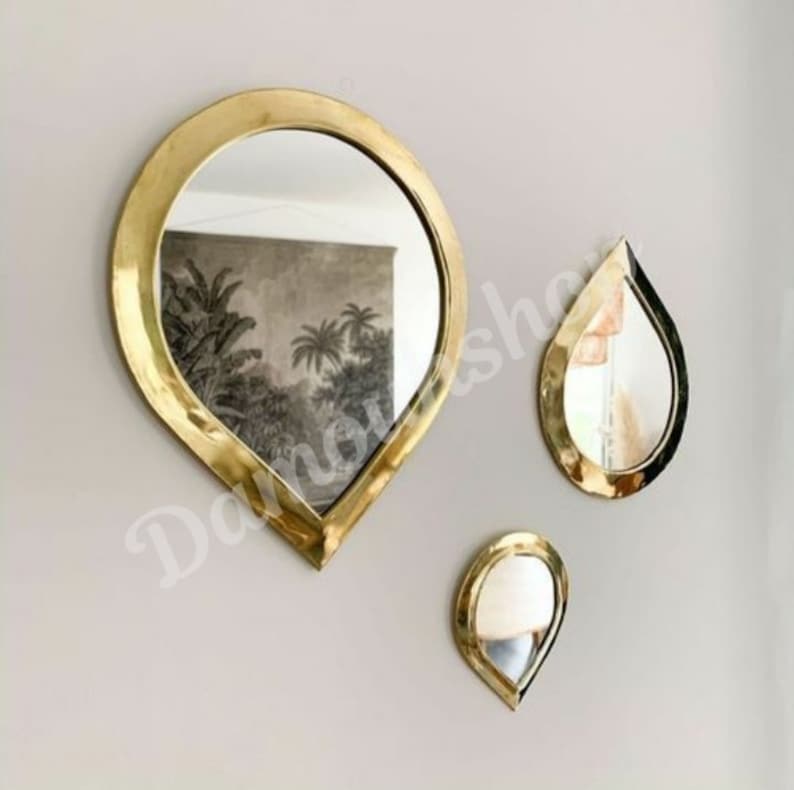 May include: Three gold teardrop-shaped mirrors of varying sizes hanging on a white wall. The largest mirror is in the centre and the two smaller mirrors are on either side.