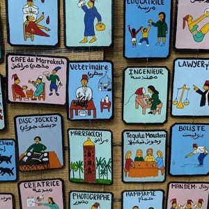 Puede incluir: Una colección de letreros coloridos pintados a mano que representan diversas profesiones. Cada letrero presenta una ilustración y texto en varios idiomas, incluyendo inglés, francés y árabe. Los letreros están dispuestos muy juntos.