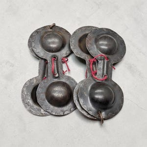 May include: A collection of antique metal finger cymbals, each with a central raised dome and decorative edge. The cymbals are connected in pairs with red string, and show signs of age and use.