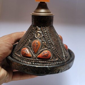 Op de afbeelding: Een decoratief, versierd, koperkleurig deksel met een conische vorm en een kleine, ronde, oranje knop bovenop. Het deksel is versierd met ingewikkelde patronen en heeft verschillende kleine, oranje, traanvormige accenten.
