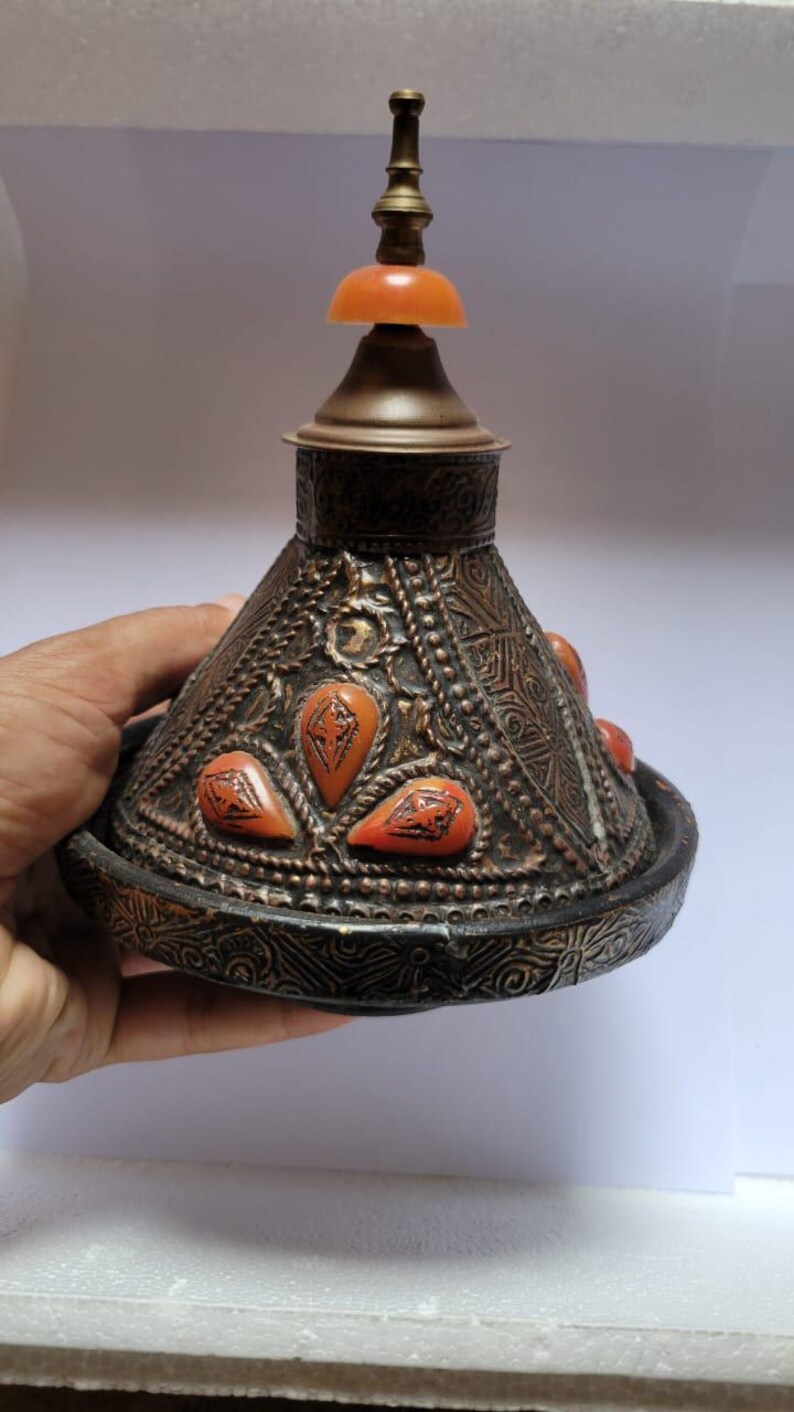 Op de afbeelding: Een decoratief, versierd, koperkleurig deksel met een conische vorm en een kleine, ronde, oranje knop bovenop. Het deksel is versierd met ingewikkelde patronen en heeft verschillende kleine, oranje, traanvormige accenten.