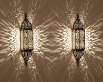 May include: Two wall-mounted lanterns featuring dark metal frames with intricate, laser-cut geometric patterns. The lanterns emit a bright, warm light that projects elaborate, starburst-like shadow patterns onto the surrounding wall surface.