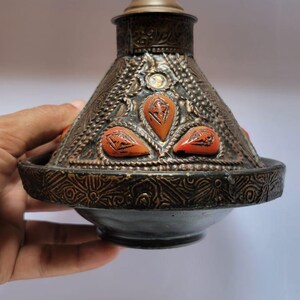 Op de afbeelding: Een decoratieve Marokkaanse tajine met een messing deksel en oranje accenten. De tajine is gemaakt van metaal en heeft ingewikkelde ontwerpen.
