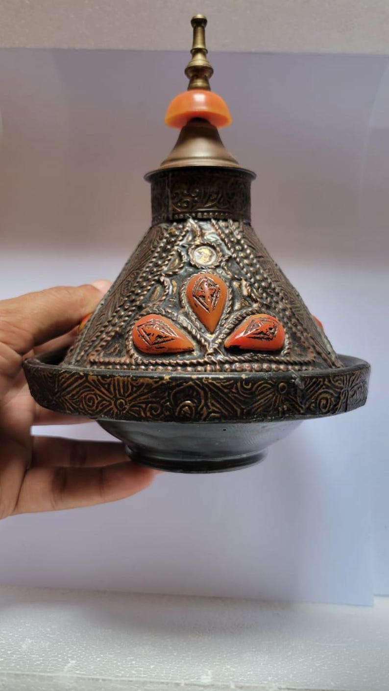 Op de afbeelding: Een decoratieve Marokkaanse tajine met een messing deksel en oranje accenten. De tajine is gemaakt van metaal en heeft ingewikkelde ontwerpen.