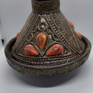 May include: A decorative, ornate, brass and copper tagine with a conical lid and a knob on top. The lid is decorated with intricate geometric patterns and has several red and orange gemstones embedded in it.