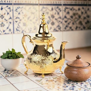 Könnte beinhalten: Eine goldfarbene, verzierte Teekanne mit passendem Deckel und kleinen Füßen. Eine kleine, runde, braune Zuckerdose mit Deckel steht neben der Teekanne. Eine kleine weiße Schale mit frischer Minze ist ebenfalls vorhanden. Die Gegenstände stehen auf einer Fliesenoberfläche.
