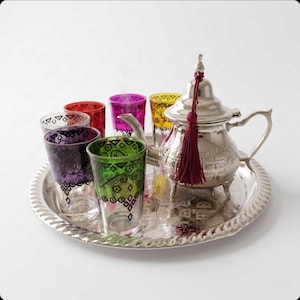 May include: A silver-toned metal teapot with a dark red tassel, accompanied by six small, colorful glass cups featuring black geometric and floral patterns, all arranged on a round, embossed silver-toned metal serving tray.