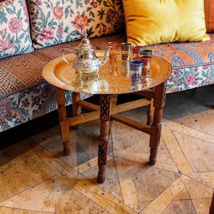 Op de afbeelding: Een kleine, ronde houten tafel met een gehamerde messing blad. Een zilveren theepot en diverse kleine, kleurrijke glazen staan op de lade. De tafel heeft gesneden poten en staat voor een bank met patronen.