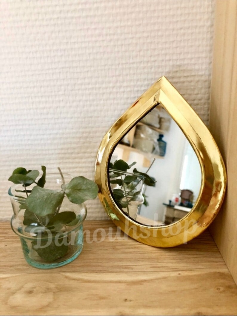 May include: A small, teardrop-shaped mirror with a gold-coloured frame. The mirror is reflecting a plant in a glass vase.