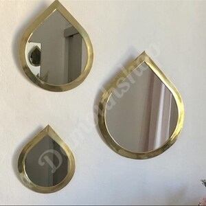 May include: Three gold teardrop-shaped mirrors of different sizes hanging on a white wall. The mirrors have a smooth, reflective surface.