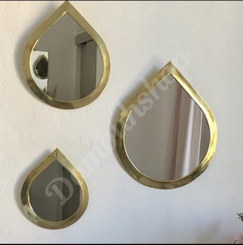 May include: Three gold teardrop-shaped mirrors of different sizes hanging on a white wall. The mirrors have a smooth, reflective surface.
