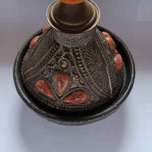 Op de afbeelding: Een zwarte keramische tajine met een decoratief oranje en gouden ontwerp. De tajine heeft een deksel met een messingknop.