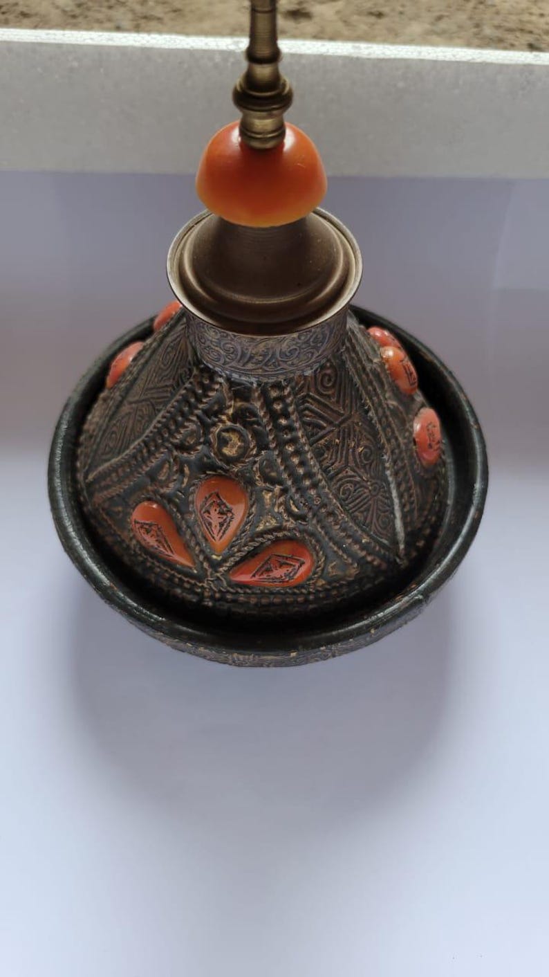 Op de afbeelding: Een zwarte keramische tajine met een decoratief oranje en gouden ontwerp. De tajine heeft een deksel met een messingknop.