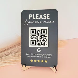 Op de afbeelding: Een rechthoekig donkergrijs bord met afgeronde hoeken, voorzien van de witte tekst PLEASE Leave us a review, een centrale QR-code, een Google G-logo en de instructie Scan this code with your phone camera to leave us a review boven vijf gouden sterren.