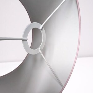 May include: A drum-shaped lampshade with a soft pink exterior and a white interior lining. The shade features a white metal spider fitter with a central ring and three support arms that connect to the inner frame of the cylindrical structure.