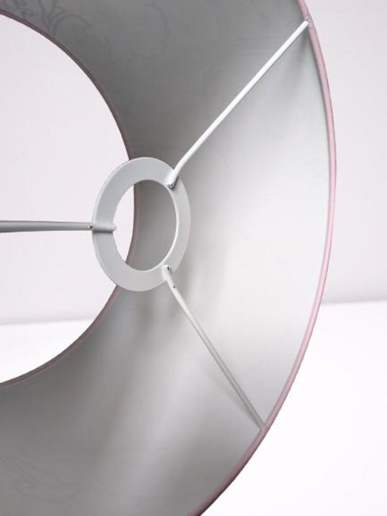 May include: A drum-shaped lampshade with a soft pink exterior and a white interior lining. The shade features a white metal spider fitter with a central ring and three support arms that connect to the inner frame of the cylindrical structure.